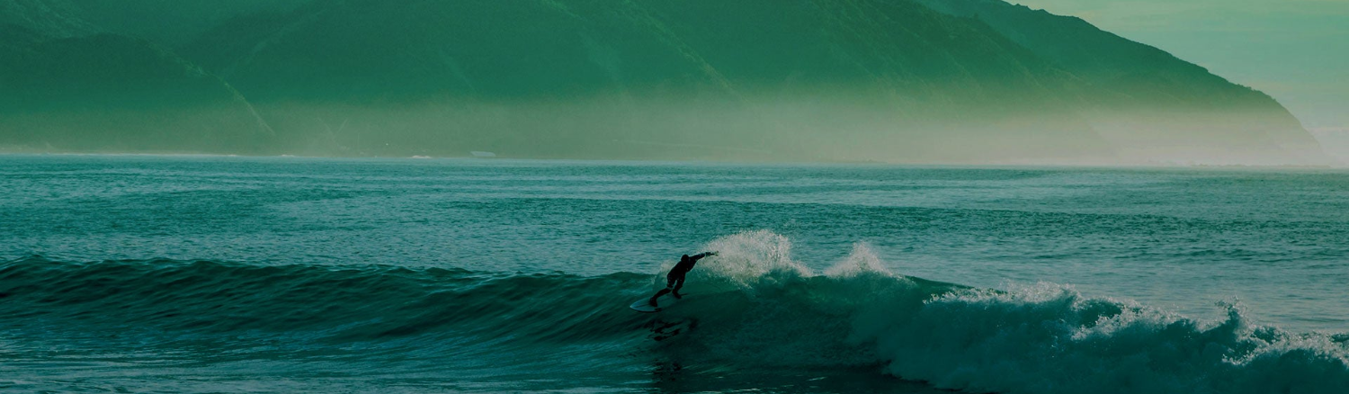 A surfer rides a wave in the ocean with a large mountain range in the background under a hazy teal sky.