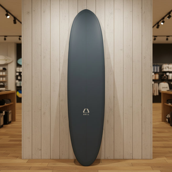 A slate blue surfboard standing upright on a yellow stand in a backyard, with a blue house, green bushes, and a paddleboard visible in the background.