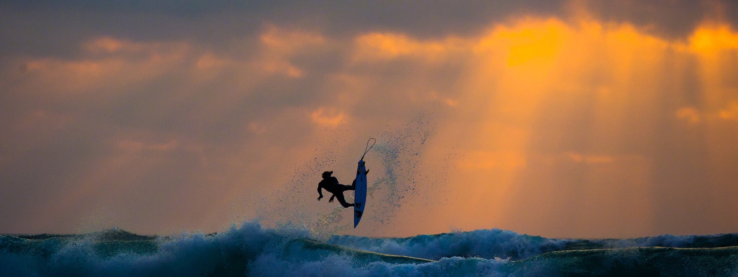 A surfer in a black wetsuit rides a board over a breaking ocean wave at sunset, with dramatic golden sun rays breaking through clouds in the sky.