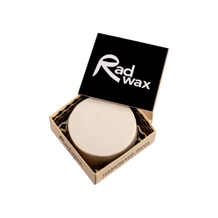 A round beige bar of Rad Wax soap in an open kraft paper box with a black lid featuring the white Rad Wax logo, set against a plain white background.