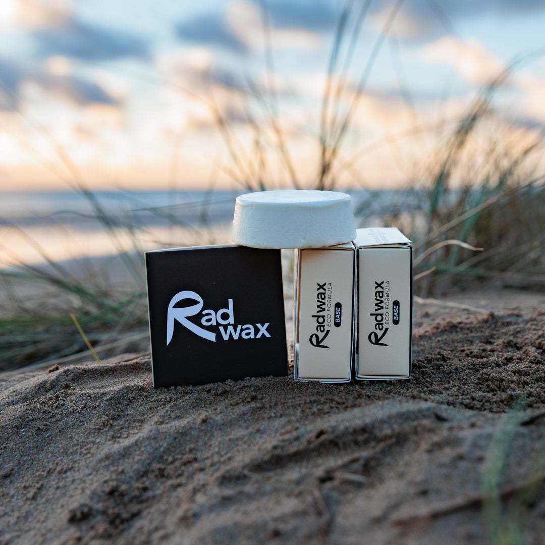 Three Radwax product boxes and a white jar are arranged on sandy dunes outdoors, with grasses and a blurred sunset sky in the background.