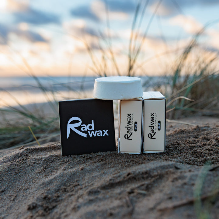 Three Radwax product boxes and a white jar are arranged on sandy dunes outdoors, with grasses and a blurred sunset sky in the background.