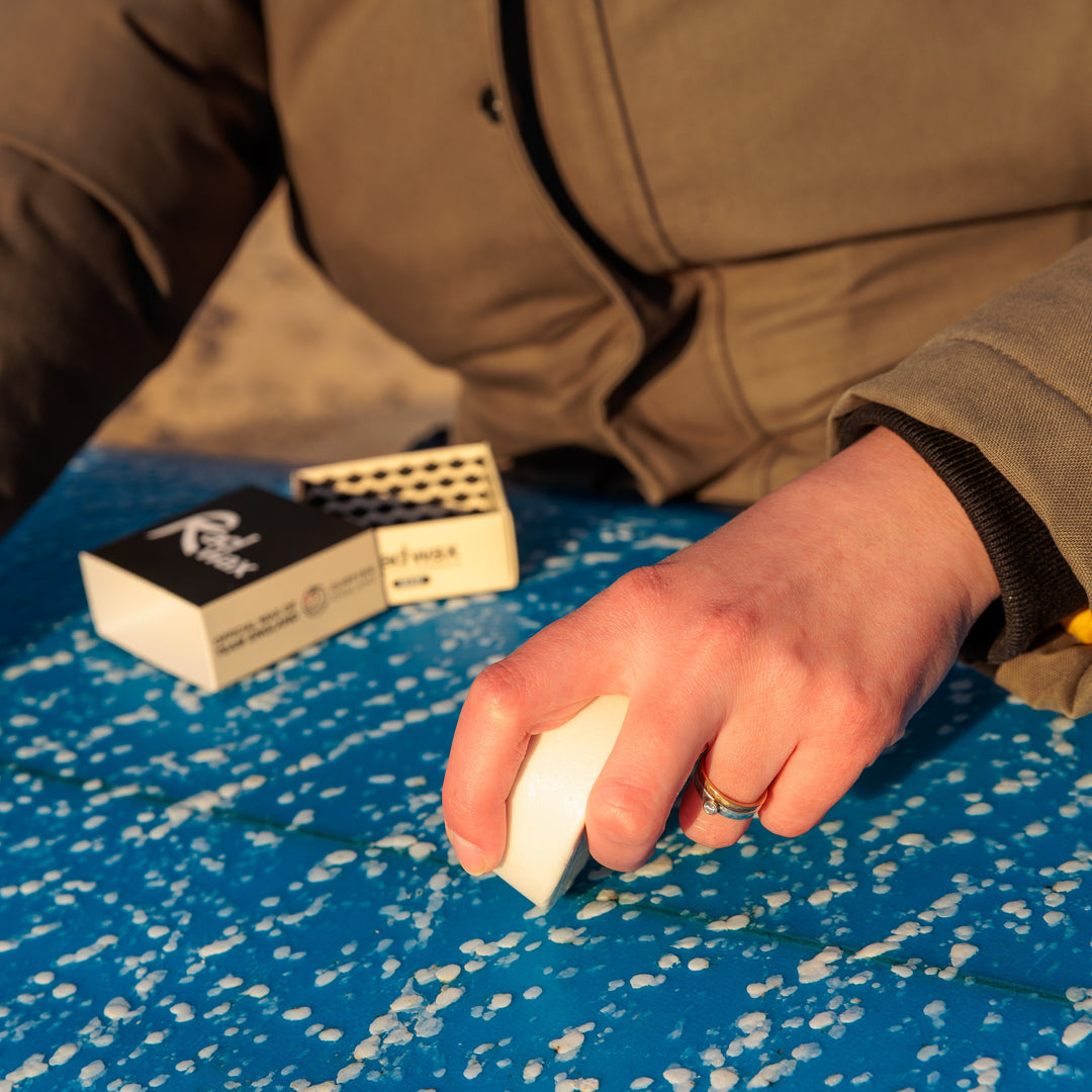 A close-up lifestyle shot of a person outdoors, wearing a brown jacket and using a white ferrocerium striker to ignite a match on a blue and white speckled blanket. A black and white matchbox is visible in the background.