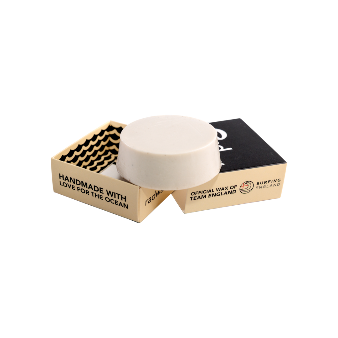 A round white wax puck displayed on top of an open cardboard surf wax box, with a second closed box behind it on a plain white background. The box is beige and black with printed text including &