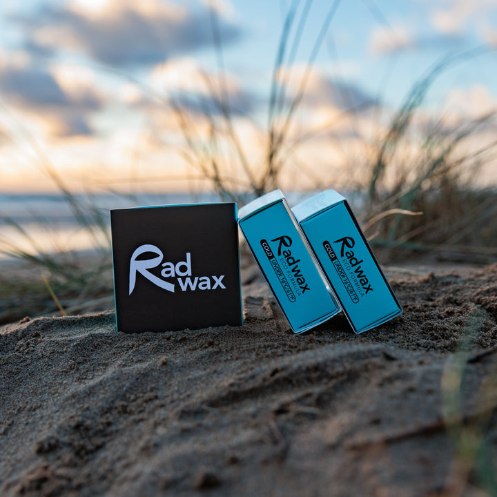 Two Radwax branded wax sticks in blue and white packaging, standing upright on sandy beach grass with a blurred ocean and sunset sky in the background.