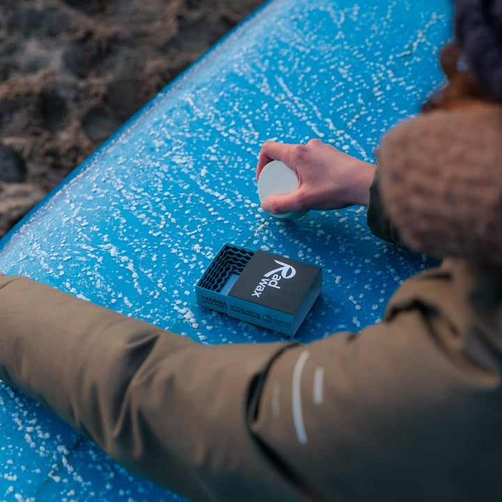 A person in a brown jacket and dark beanie holds a small white circular item over a bright blue surfboard with a black branded case on it, outdoors near dark sand.