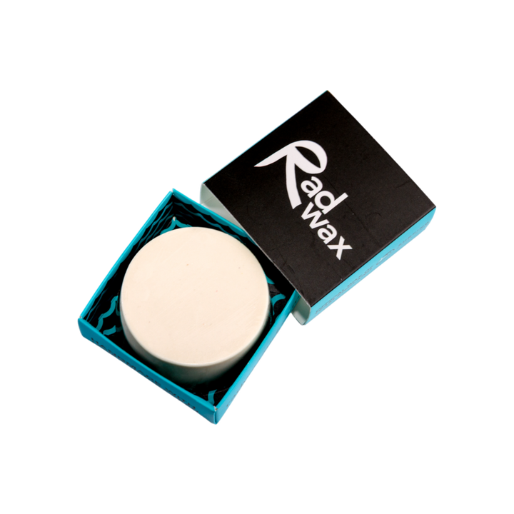 A round white wax melt or solid stick product displayed inside an open teal and black box, with the lid showing the white &