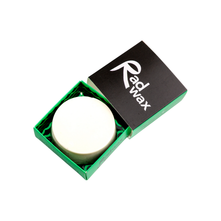 A round white wax melt or solid bar product presented in an open green box with a black lid featuring the white &