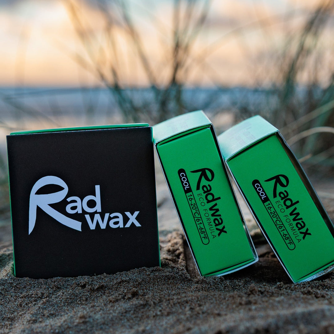 Three Radwax cooling wax boxes on a sandy beach with blurred grass and water in the background. One box is black with a white logo, and two boxes are bright green with black and white text.