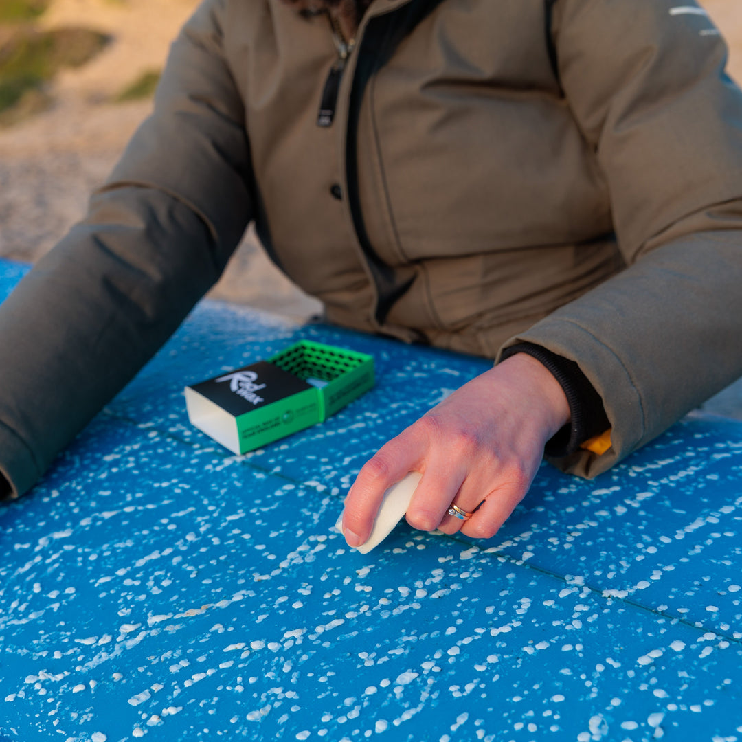 A close-up of a person outdoors applying white surf foam from a small white squeeze bottle onto a bright blue surfboard. A small green and black surf wax container sits nearby on the board.