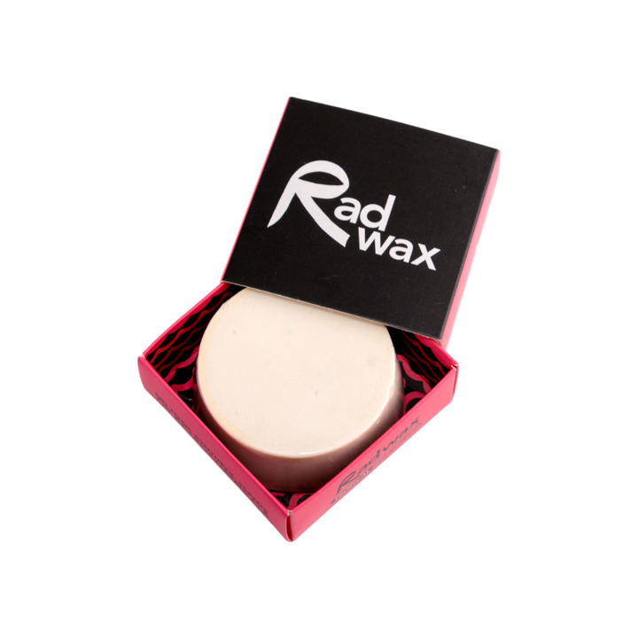 A round beige bar of wax presented in an open pink and black Rad Wax gift box, photographed on a solid white background.