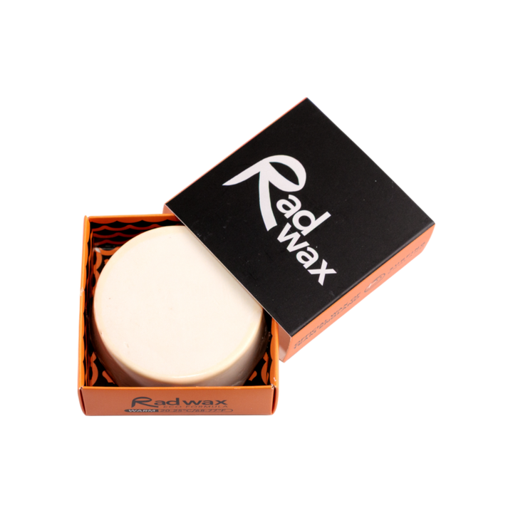 A round white bar of Radwax body wax displayed in its orange cardboard box with a black lid featuring the white Radwax logo, set against a plain white background.