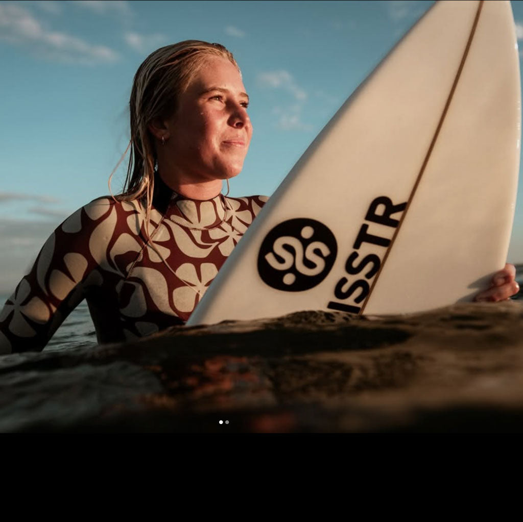 A woman in a patterned wetsuit holding a white surfboard with a black circular 'SS' logo and the word 'ISSTR' near the bottom, photographed outdoors by the water under a blue sky.
