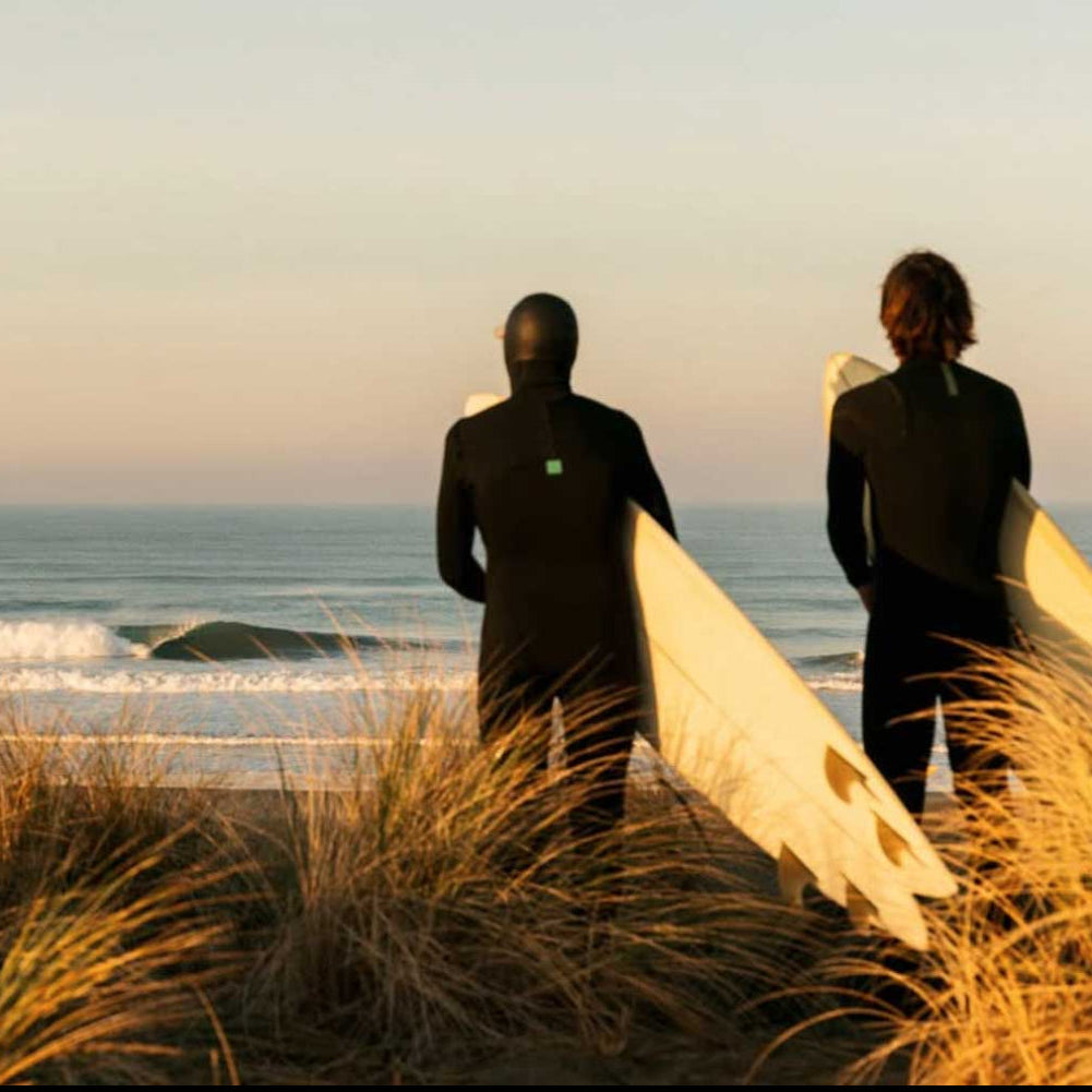 Two surfers in black wetsuits standing on a beach dune at sunset, holding white surfboards and looking out toward the ocean.