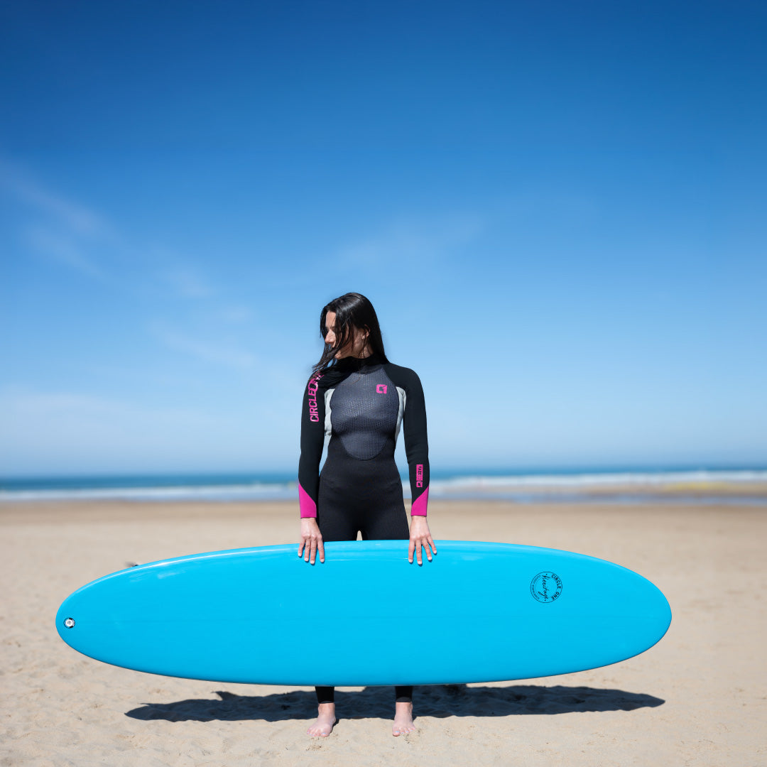 A woman stands on a sandy beach holding a bright blue surfboard, wearing a black Delta wetsuit with pink accents against a clear blue sky and ocean background.