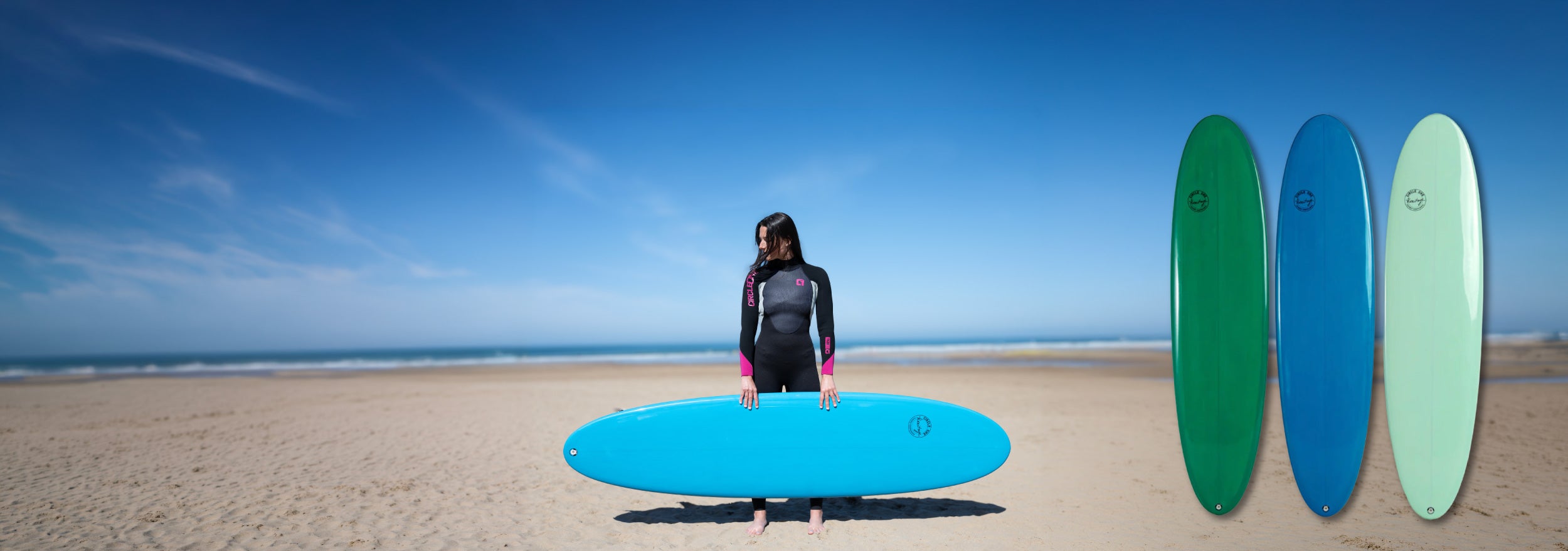 A person in a black wetsuit stands on a sandy beach holding a bright blue surfboard. Three additional surfboards in green, blue, and mint are shown vertically on the right side against the beach and blue sky background.